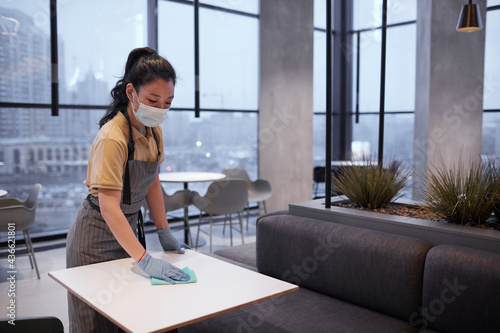 Portrait of young female worker wearing mask while cleaning tables in restaurant or food court interior, copy space