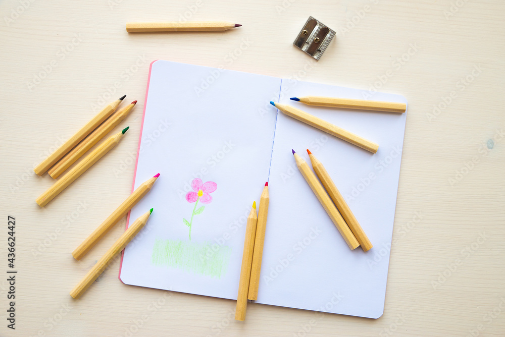 Open white notebook, with colored crayons beside it, penknife and drawing of a colored flower. Open white notebook, with colored crayons beside it, penknife and drawing of a colored flower.