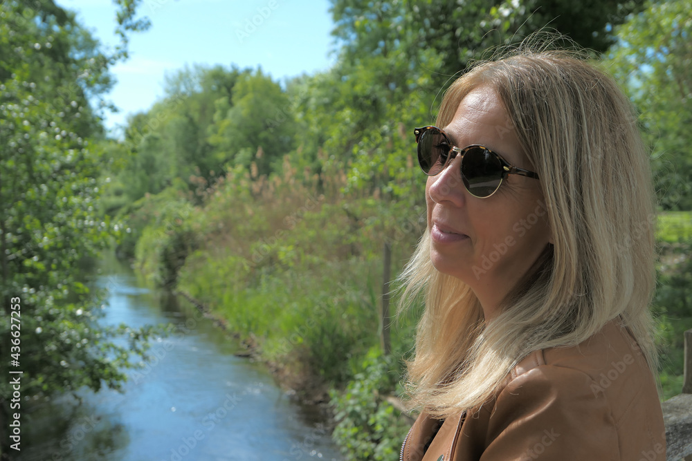 Beautiful blonde woman with long blonde hair, in profile, at the water's edge in nature. Lake and vegetation blurred voluntarily in the background. Beautiful blonde woman with long blonde hair, in profile, at the water's edge in nature. Lake and vegetation blurred voluntarily in the background.