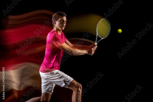 Young caucasian male tennis player playing tennis in mixed light on dark background. Concept of healthy lifestyle, professional sport, hobby. © master1305