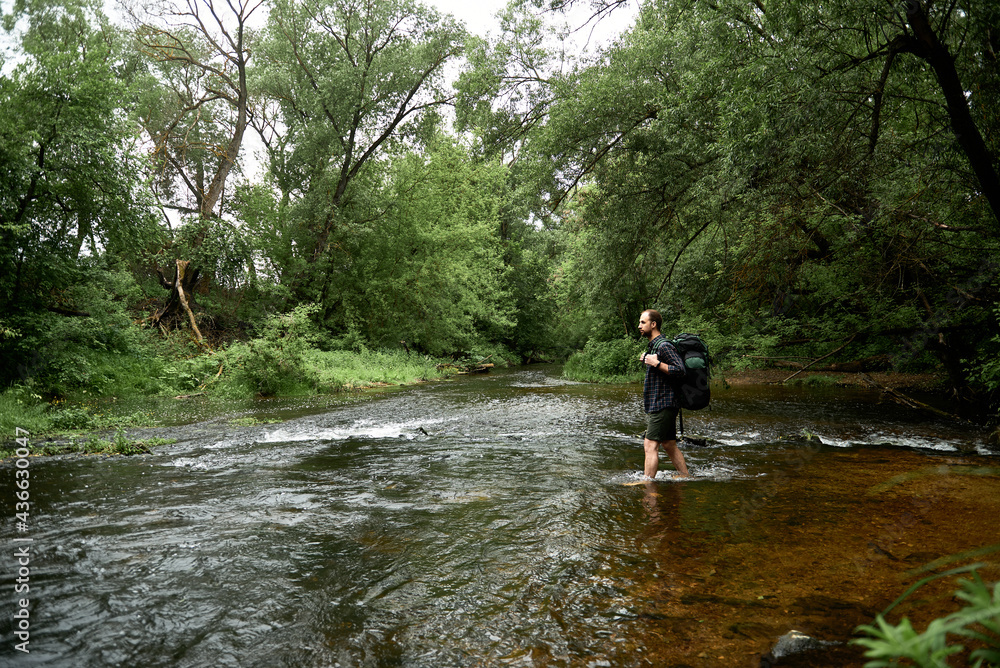A man in a plaid shirt and green shorts with a large hiking backpack crosses the river