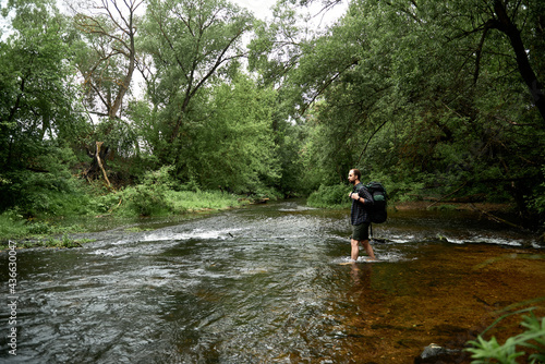 A man in a plaid shirt and green shorts with a large hiking backpack crosses the river