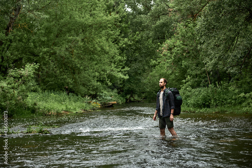 A man in a plaid shirt and green shorts with a large hiking backpack crosses the river