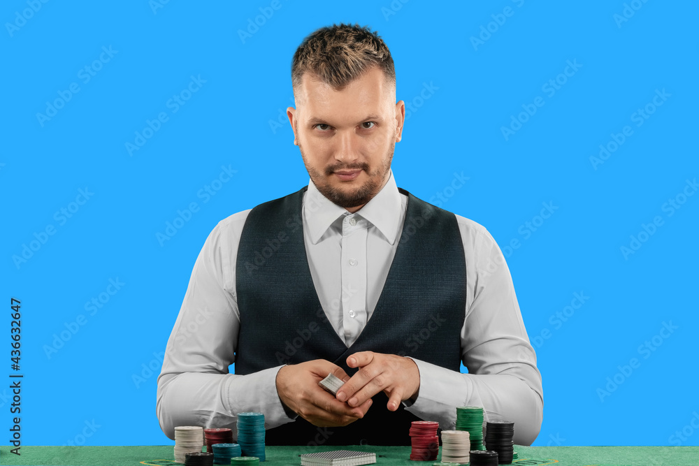 Male croupier at the casino at the table isolated on blue background. Casino concept, gambling, poker, chips on the green casino table. Male croupier at the casino at the table isolated on blue background. Casino concept, gambling, poker, chips on the green casino table.