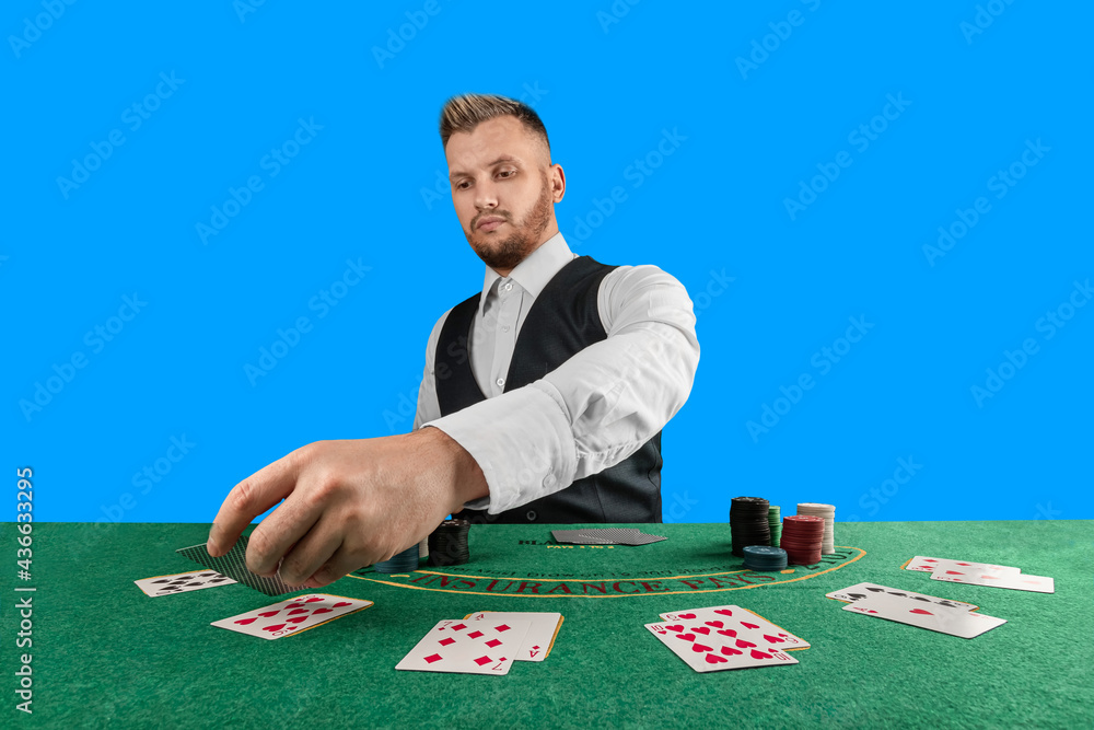 Male croupier at the casino at the table isolated on blue background. Casino concept, gambling, poker, chips on the green casino table. - obrazy, fototapety, plakaty 