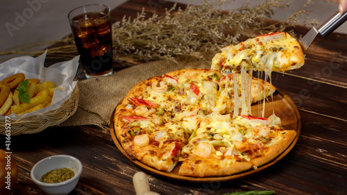 Selective focus and close up on a piece of double cheese homemade seafood pizza on spatula or scoop with blur background of wooden table, sackcloth, ingredients and fried potatoes. Food Concept.
