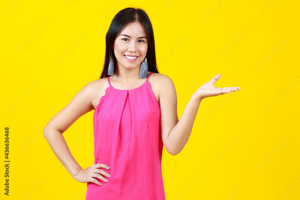 Asian beautiful happy cheerful adult woman with long black hair wearing pink dress, fashionable earring, showing hand present good or product, isolated on yellow background. Asian beautiful happy cheerful adult woman with long black hair wearing pink dress, fashionable earring, showing hand present good or product, isolated on yellow background.