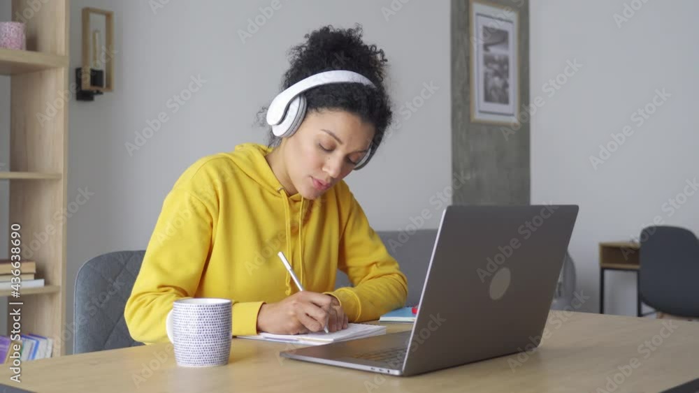 African American mixed race adult student wearing headphones having virtual meeting online class with teacher, writing notes. Webinar chat at home office. Video e learning conference call on pc. African American mixed race adult student wearing headphones having virtual meeting online class with teacher, writing notes. Webinar chat at home office. Video e learning conference call on pc.