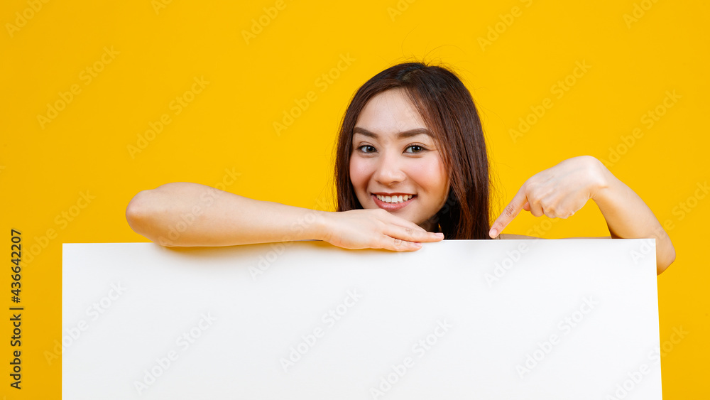 Cute and pretty curly hair Asian female brunette holding and pointing at white blank board poses to camera with a joyful for advertising and banner use purpose, isolated on bright yellow background Cute and pretty curly hair Asian female brunette holding and pointing at white blank board poses to camera with a joyful for advertising and banner use purpose, isolated on bright yellow background