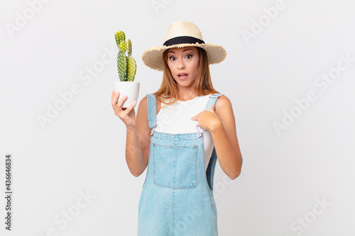 pretty woman looking shocked and surprised with mouth wide open, pointing to self and holding a cactus decorative plant photo