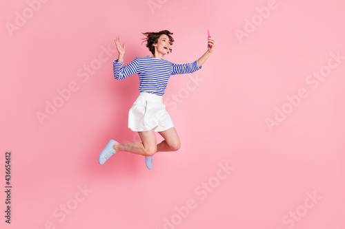 Full size photo of young beautiful excited happy cheerful girl take selfie say hi jump isolated on pink color background