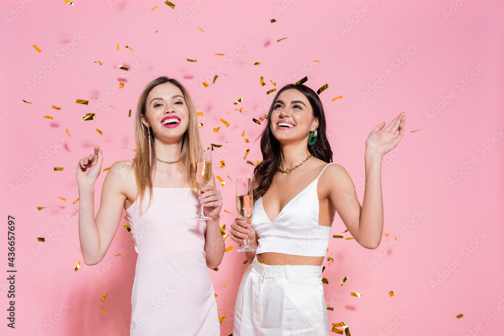 Positive brunette woman with champagne standing near friend and confetti on pink background.