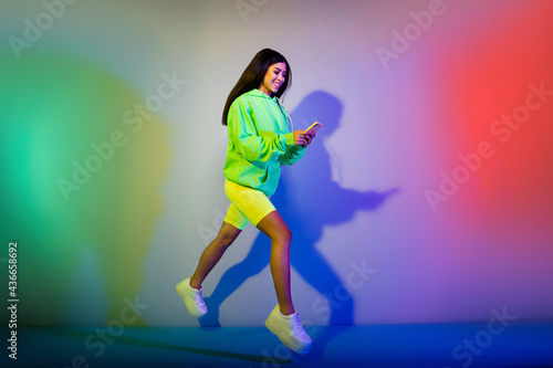 Full length body size view of beautiful trendy girl jumping using gadget chatting isolated over multicolor vivid neon light color background