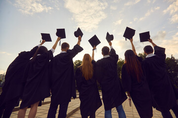 Youth having high hopes. Back view of group of university graduates standing in row holding up black academic caps. Confident college students together tossing hats in air at graduation party