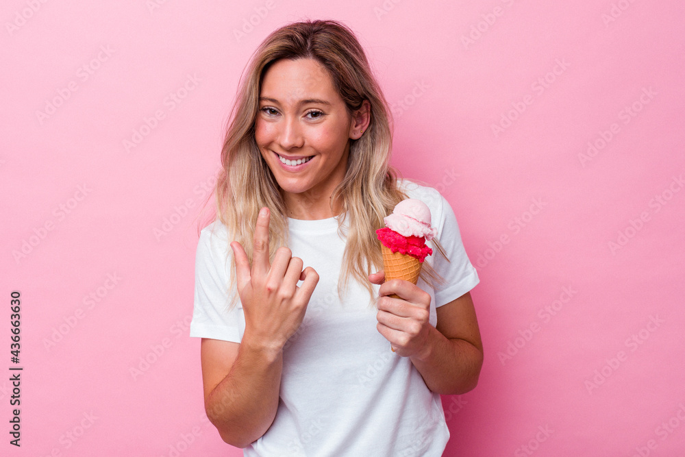 Young australian woman holding an ice cream isolated on pink background pointing with finger at you as if inviting come closer.