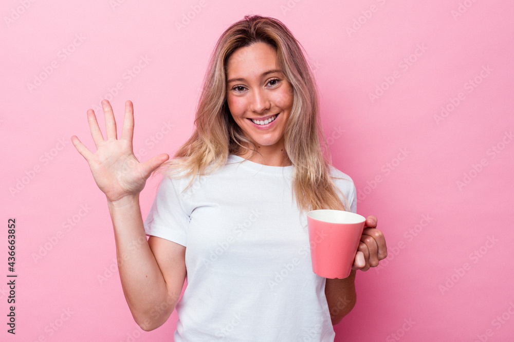 Young australian woman holding a pink mug isolated on pink background smiling cheerful showing number five with fingers. Young australian woman holding a pink mug isolated on pink background smiling cheerful showing number five with fingers.