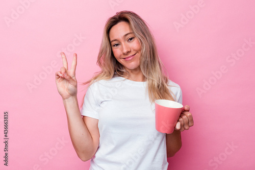 Young australian woman holding a pink mug isolated on pink background showing number two with fingers.