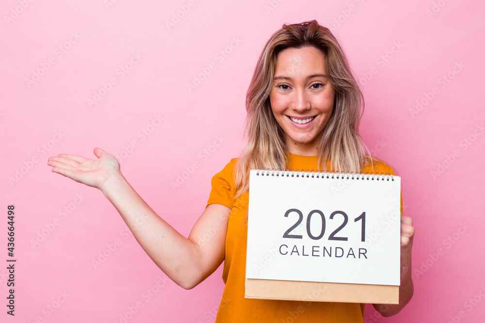 Young australian woman holding a calendar isolated on pink background showing a copy space on a palm and holding another hand on waist. Young australian woman holding a calendar isolated on pink background showing a copy space on a palm and holding another hand on waist.