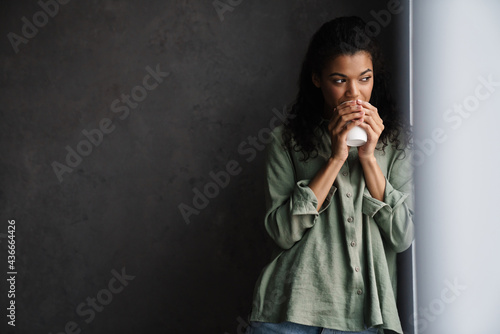 Fotografía Black young woman drinking coffee while leaning on wall indoors