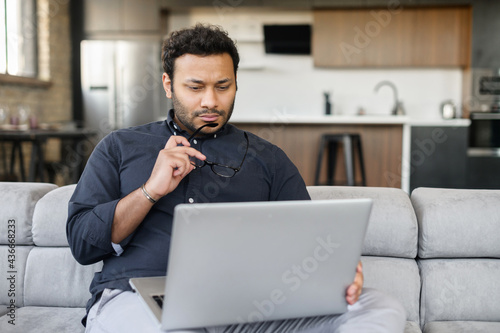 Thoughtful indian man using laptop sitting on the couch at home, a hindu guy looks at the screen, looking for new sturtup idea, solving difficalt tasks, planning future photo