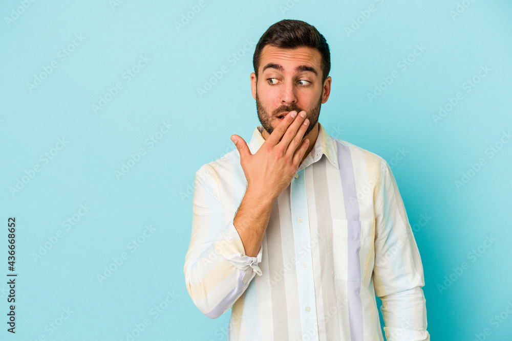 Young caucasian man isolated on blue background thoughtful looking to a copy space covering mouth with hand. Young caucasian man isolated on blue background thoughtful looking to a copy space covering mouth with hand.