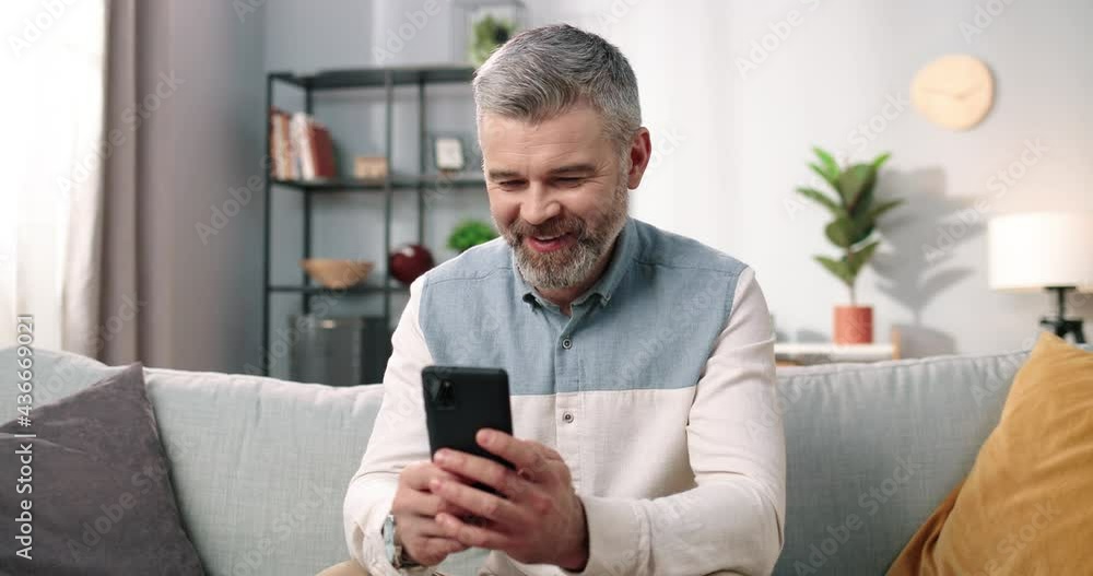 Close up of cheerful handsome middle-aged Caucasian man in positive mood texting on smartphone while sitting at modern home on sofa and smiling, grey-haired male using social network app on cellphone Close up of cheerful handsome middle-aged Caucasian man in positive mood texting on smartphone while sitting at modern home on sofa and smiling, grey-haired male using social network app on cellphone