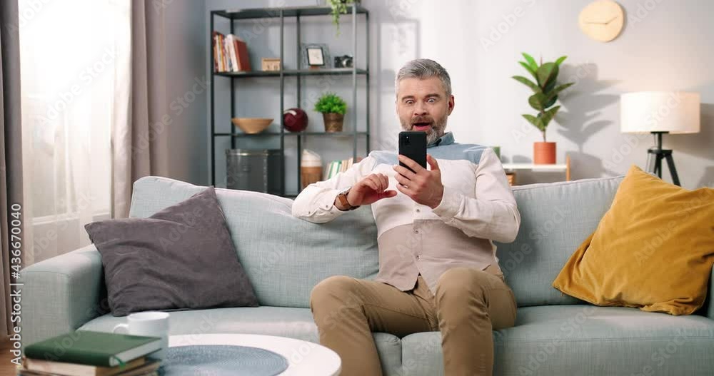 Happy Caucasian handsome middle-aged bearded man in good mood texting on smartphone with amazed shocked face while sitting at home on sofa, grey-haired male using social network app on cellphone Happy Caucasian handsome middle-aged bearded man in good mood texting on smartphone with amazed shocked face while sitting at home on sofa, grey-haired male using social network app on cellphone