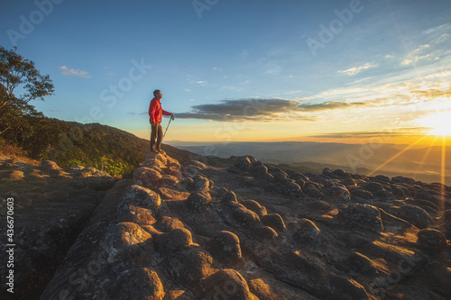 One man in a red jacket with a trekking stick is standing on a rocky hilltop. Sunset time There is a beautiful sky.