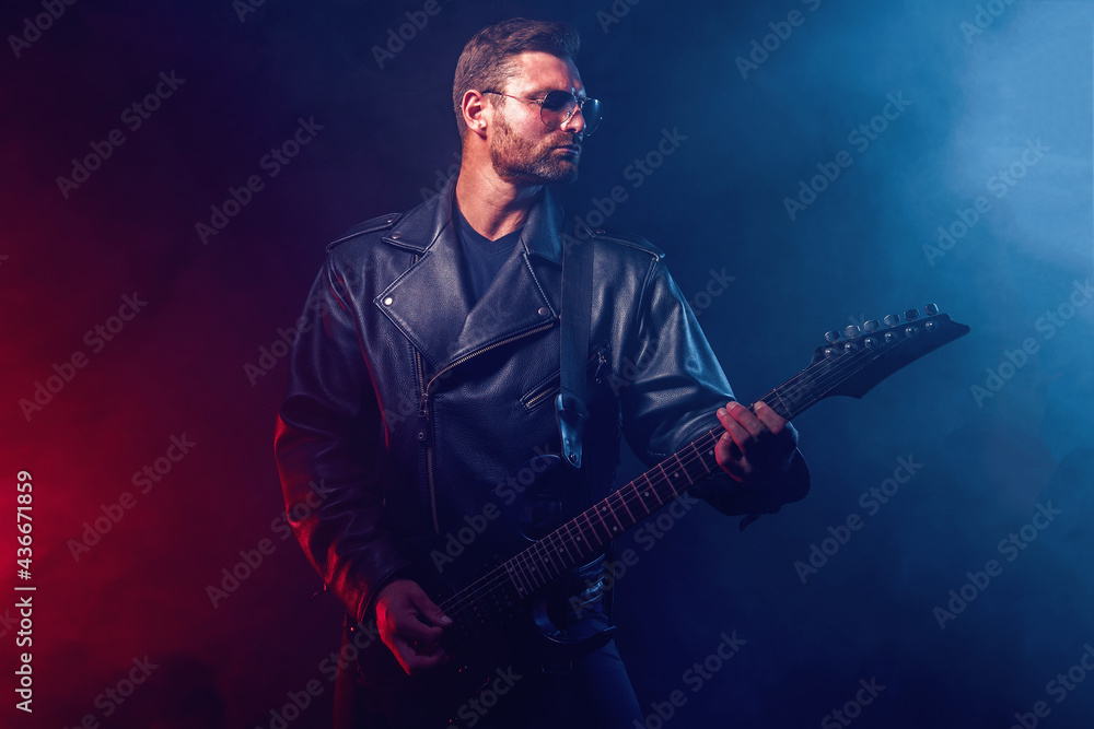 Obraz premium Brutal bearded Heavy metal musician in leather jacket, cap and sunglasses is playing electrical guitar. Shot in a studio on dark background