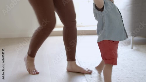 Medium shot of happy boy taking his first steps with help of mom. Curly-haired boy holding mothers hand, walking on carpet. First steps, family concept. photo