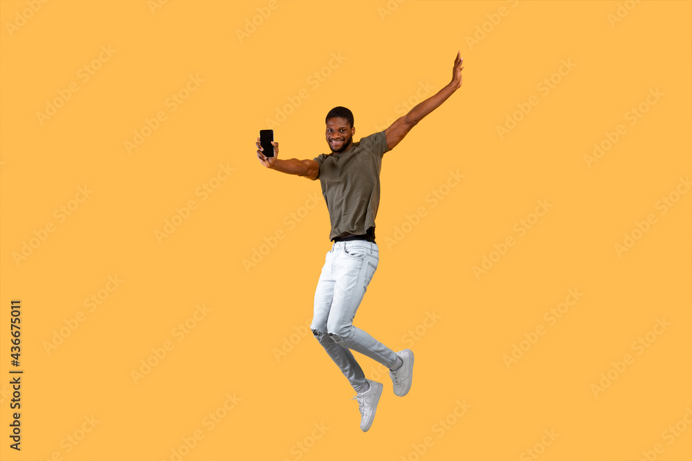 Joyful african americna man jumping over yellow background and showing smartphone with black screen to camera.