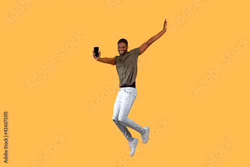 Joyful african americna man jumping over yellow background and showing smartphone with black screen to camera.