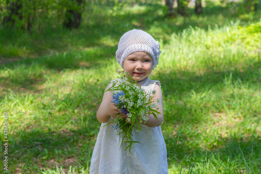 Cute baby girl in white dress and panama hat in summer holds a bouquet of delicate wildflowers in the forest