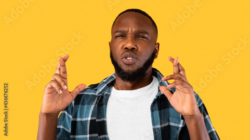 Black Guy Keeping Fingers Crossed Frowning Standing Over Yellow Background