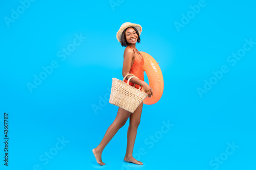 Summer beach vacation. Full length portrait of black woman in bikini going to pool with inflatable ring and straw bag © Prostock-studio