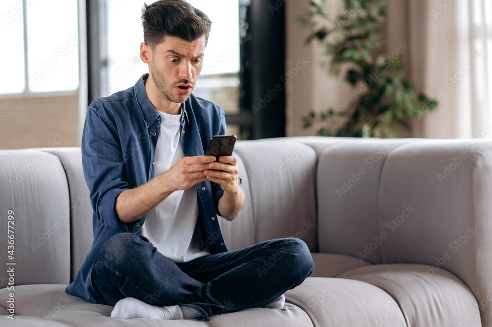 Obraz premium Shocked confused young caucasian man, in casual wear, sitting on sofa in lotus position, uses smart phone, receiving an unexpected message, texting with friends, looking at screen in surprise