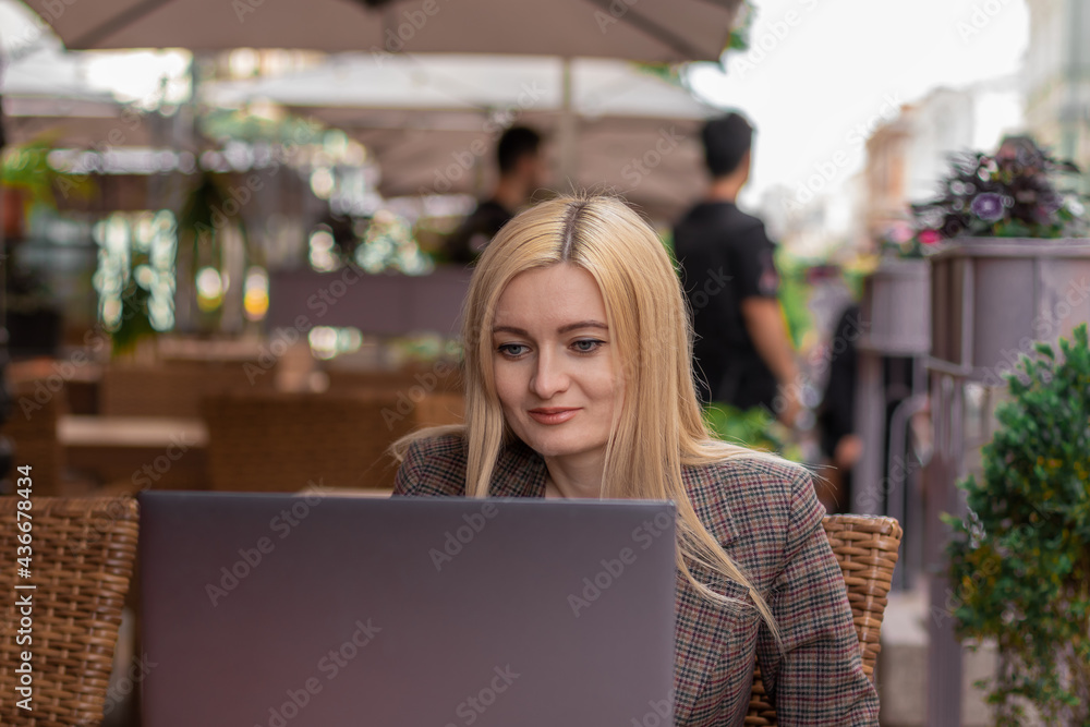 young blonde woman sitting with laptop on summer terrace and smiling. front view