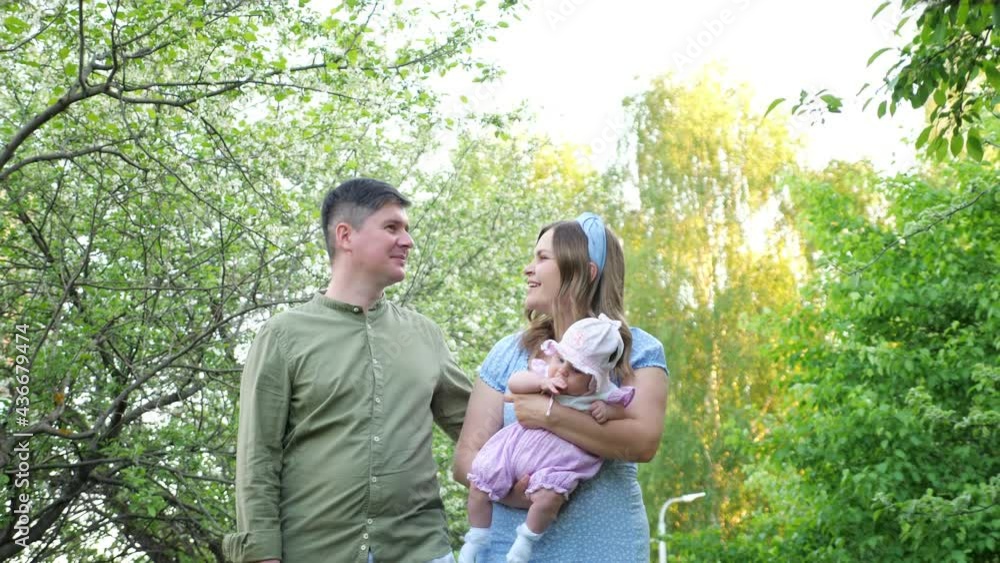 Joyful family husband and wife wanders holding in arms baby daughter along green park with white blossom and smiles on spring day slow motion Joyful family husband and wife wanders holding in arms baby daughter along green park with white blossom and smiles on spring day slow motion