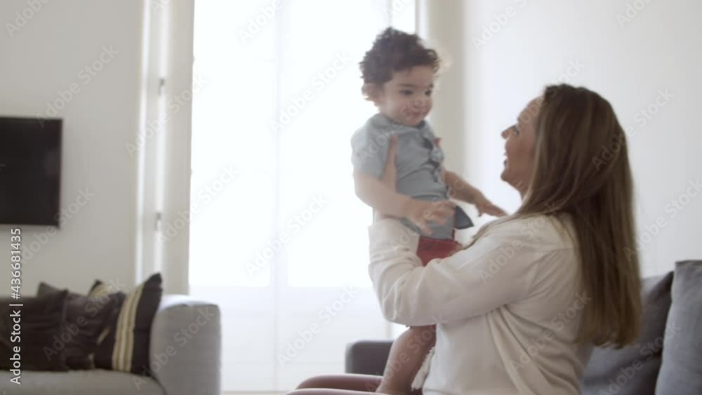 Focused boy taking first steps, walking to his mom. Excited Caucasian mother in casual wear sitting on sofa in living room, lifting her little son. First steps, motherhood concept. Focused boy taking first steps, walking to his mom. Excited Caucasian mother in casual wear sitting on sofa in living room, lifting her little son. First steps, motherhood concept.
