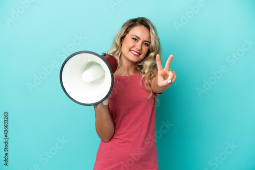 Young Brazilian woman isolated on blue background holding a megaphone and smiling and showing victory sign