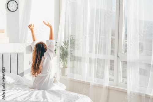  Woman in white bathrobe sitting near the big white window while stretching on bed after waking up.