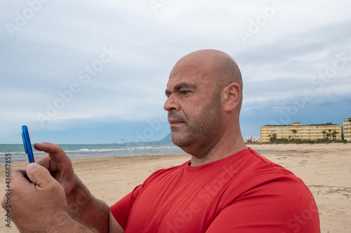 Stocky Caucasian middle-aged man using mobile phone to send messages on an empty beach at sunset. photo