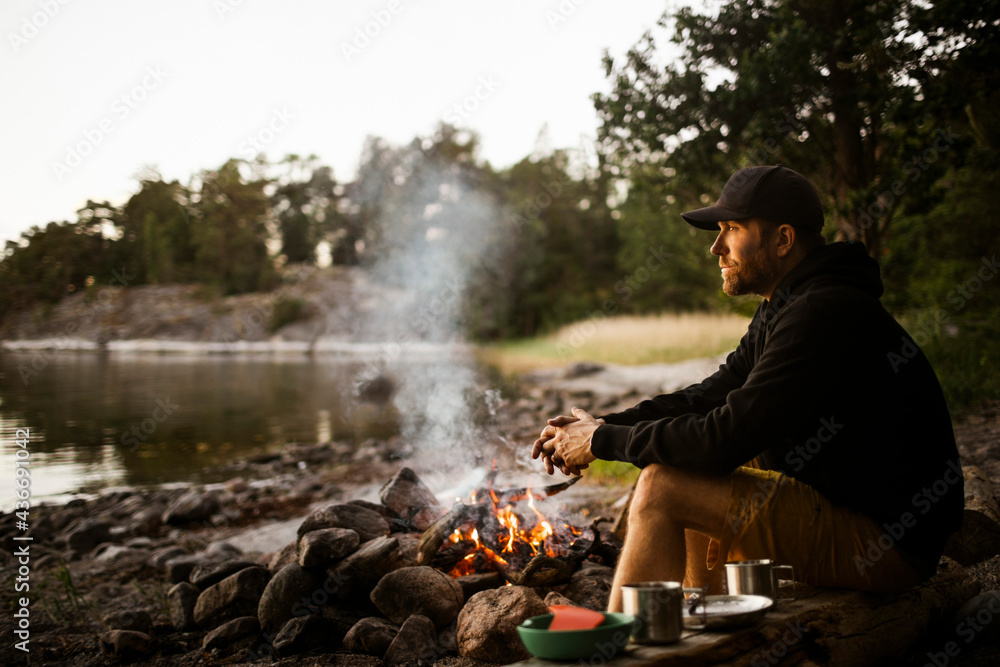 Side view of thoughtful man looking away while sitting by campfire at lakeshore Side view of thoughtful man looking away while sitting by campfire at lakeshore