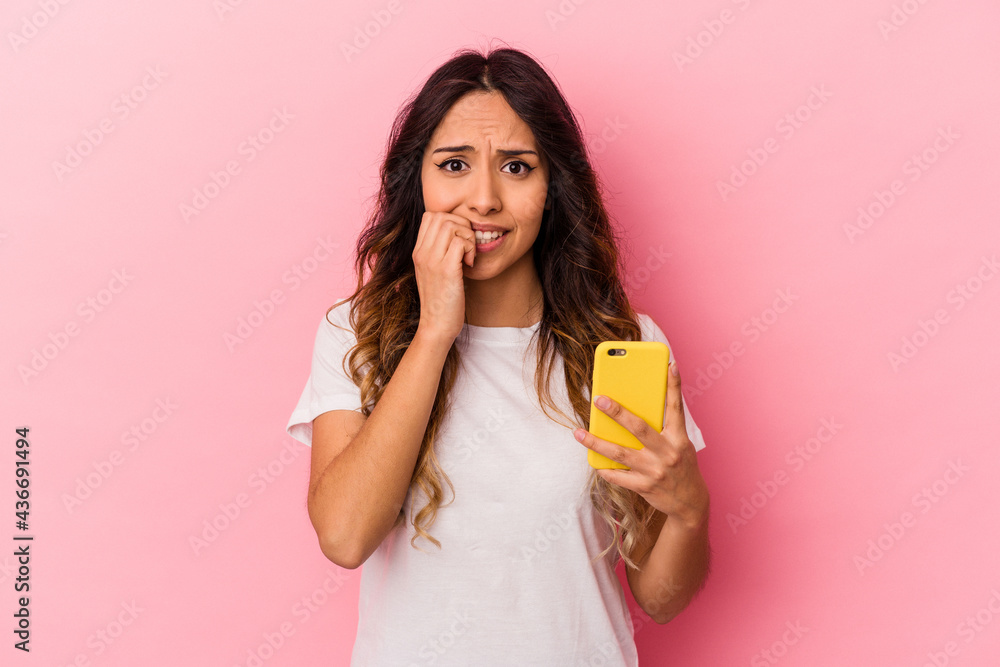 Young mexican woman holding a mobile phone isolated on pink background biting fingernails, nervous and very anxious. Young mexican woman holding a mobile phone isolated on pink background biting fingernails, nervous and very anxious.