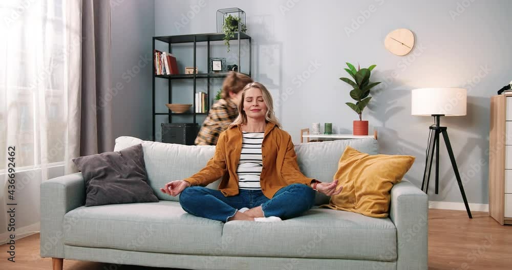 Beautiful positive Caucasian mother meditating with calm face and closed eyes in living room on sofa while her little son child screaming running around playing, family concept, yoga meditation Beautiful positive Caucasian mother meditating with calm face and closed eyes in living room on sofa while her little son child screaming running around playing, family concept, yoga meditation