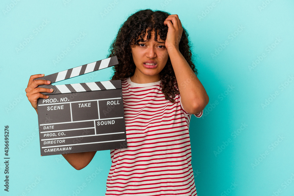 Young mixed race woman holding a clapperboard isolated on blue background being shocked, she has remembered important meeting.
