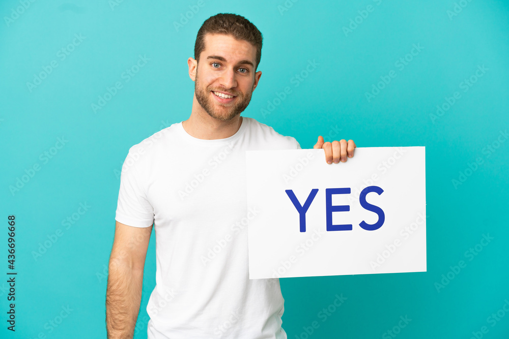 Handsome blonde man over isolated blue background holding a placard with text YES with happy expression Handsome blonde man over isolated blue background holding a placard with text YES with happy expression