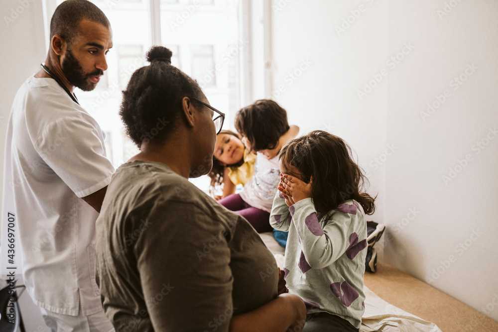 Male pediatrician and woman looking at girl covering eyes sitting with playful sisters Male pediatrician and woman looking at girl covering eyes sitting with playful sisters