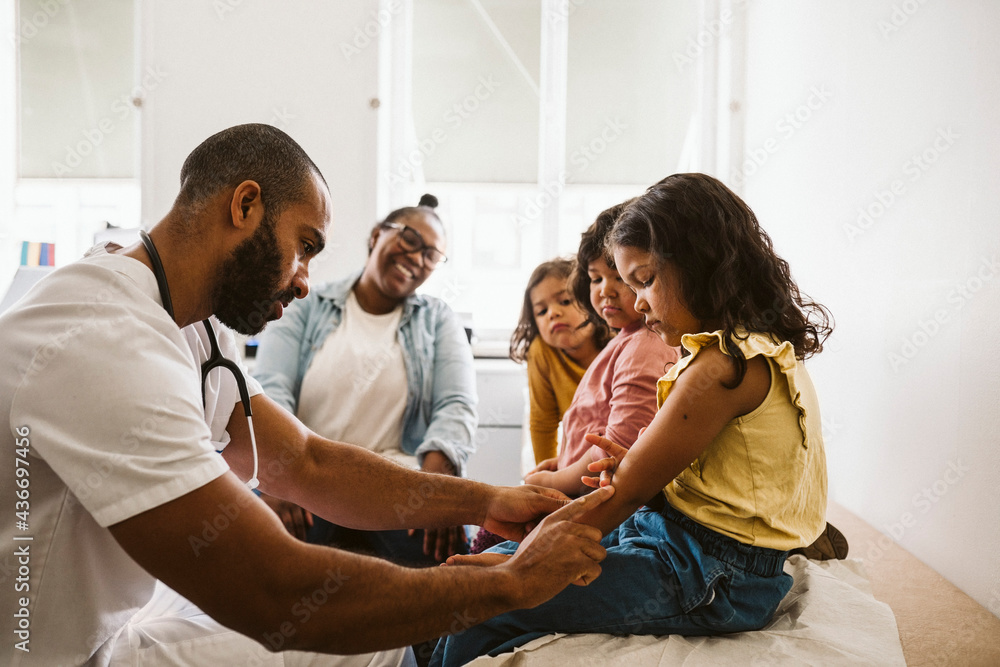 Mother and daughters looking at male doctor examining girl's hand at medical clinic Mother and daughters looking at male doctor examining girl's hand at medical clinic