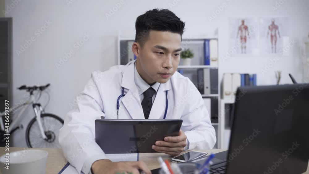 asian man doctor is referring to the laptop while studying a patient’s file on the electronic pad with finger pointing and following the data in his office. asian man doctor is referring to the laptop while studying a patient’s file on the electronic pad with finger pointing and following the data in his office.